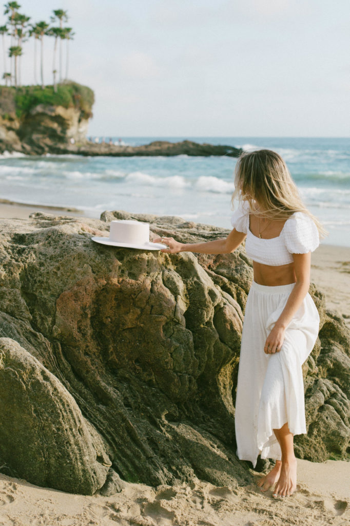 model is reaching for white hat on top of rock at the beach