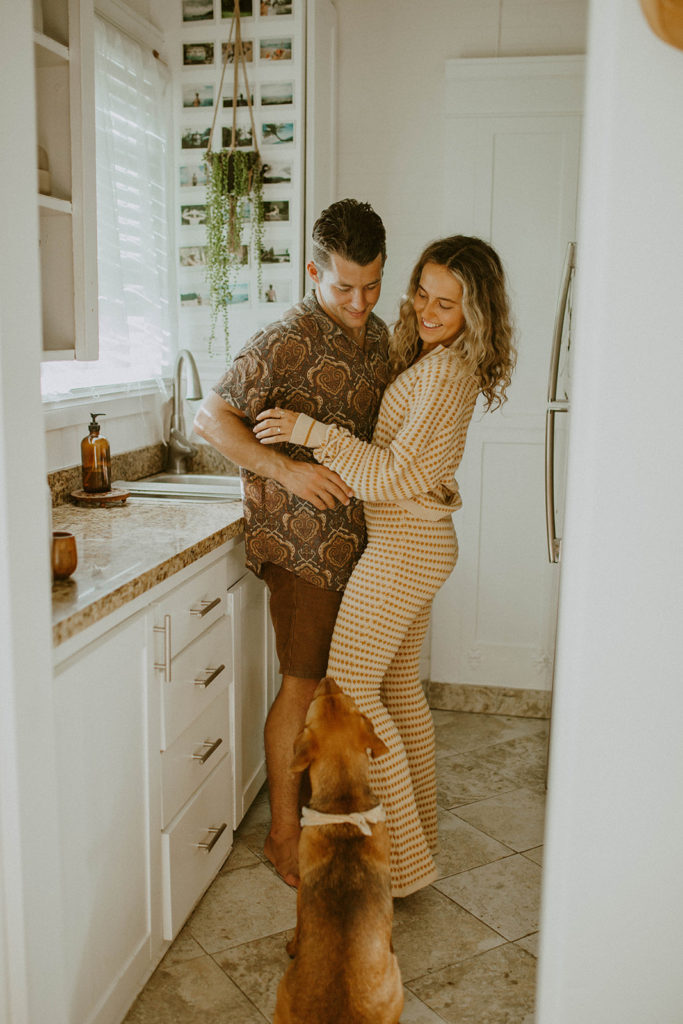 Couple is in kitchen hugging each other while looking at their dog