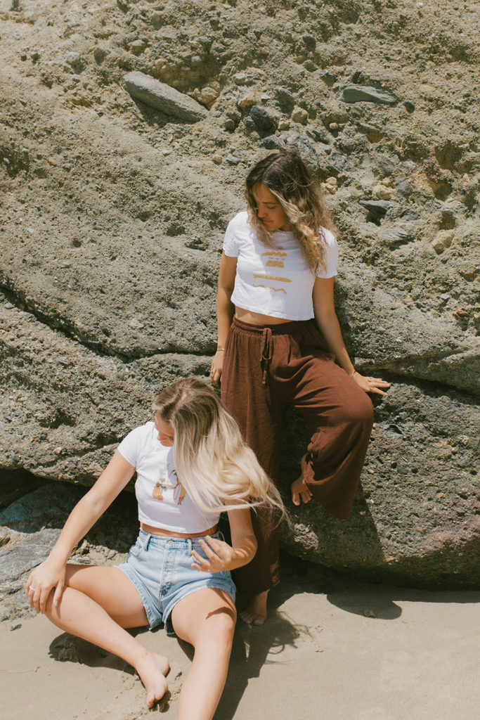 both models are leaning against a rock on the beach and both wearing saltee hawaii graphic tee's