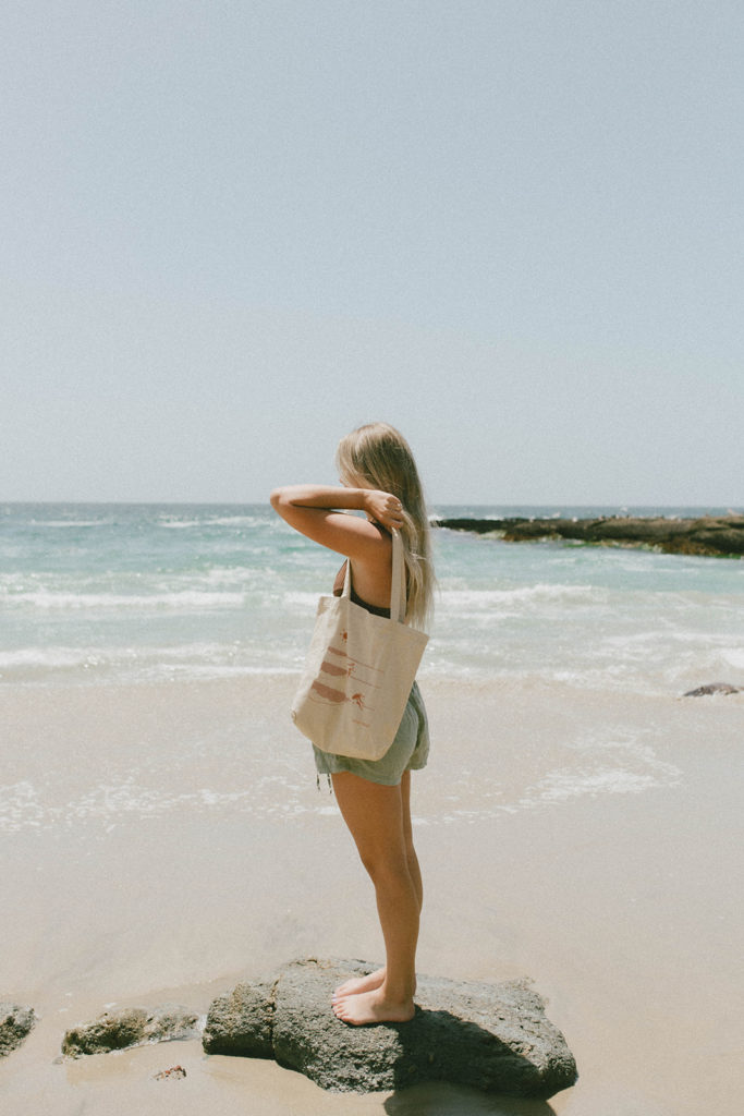 model has saltee hawaii's graphic tote draped across her shoulder while standing on rock on the beach