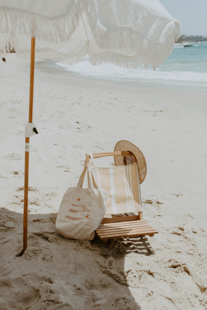 picture of beach chair with hat and bag hanging from it, while under an umbrella at the beach