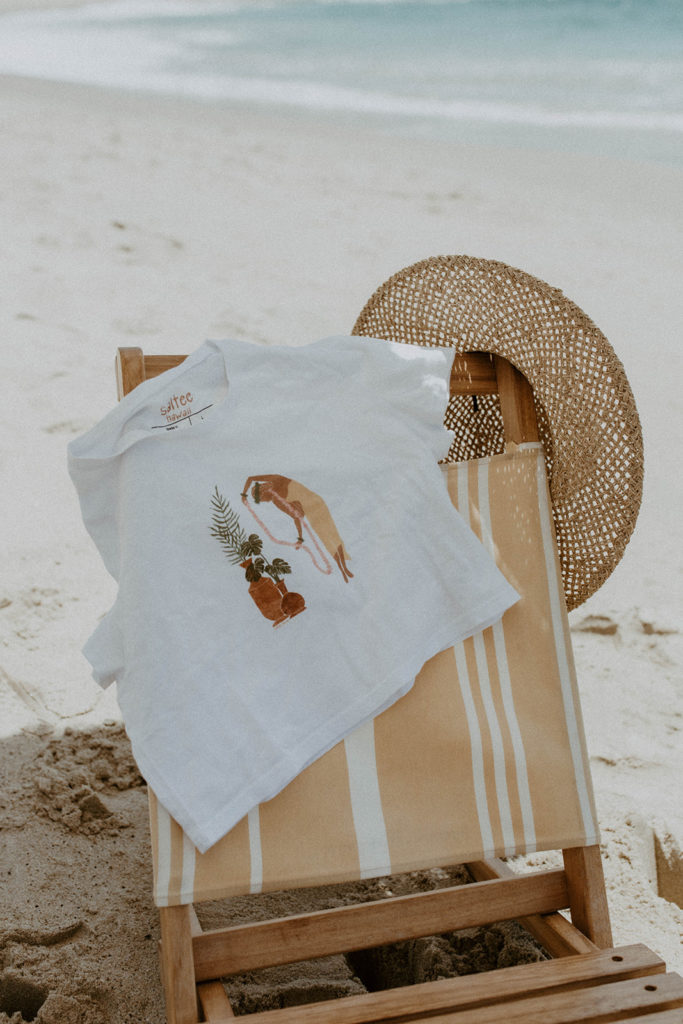picture of beach chair with saltee hawaii's graphic tee draped across it with a beach hat placed on chair too