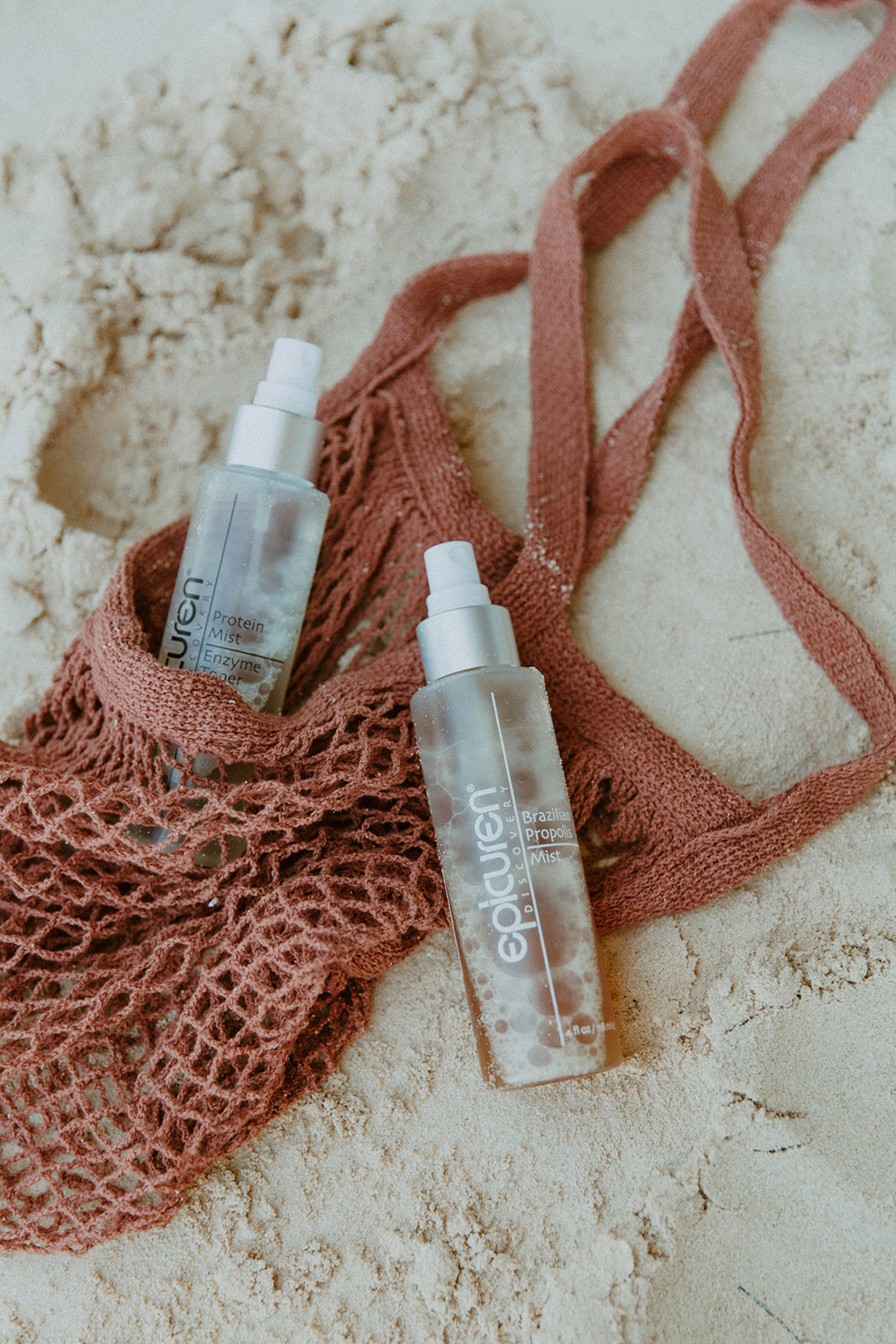 shot of two products in the sand by a pink woven beach bag